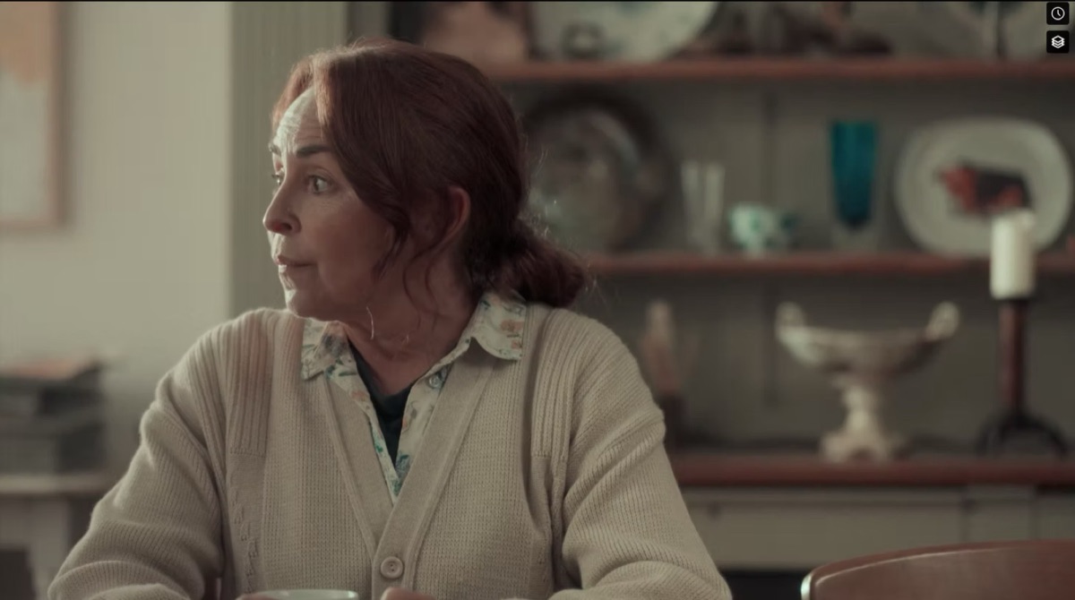 Still from Something Else: a woman in a cream cardigan at a table in a lived-in kitchen.