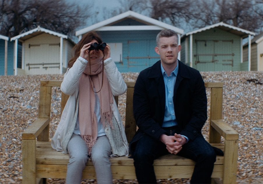Still from The Sea: Anna Friel looking through binoculars beside Russell Tovey on a wooden bench in front of beach huts.