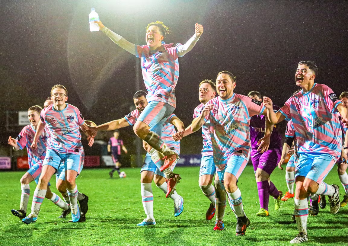 Still from We'll Go Down in History: footballers in pink and blue kits celebrating on a floodlit pitch in the rain.
